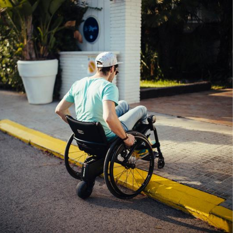 Homem numa cadeira de rodas equipada com o Alber Smoov One, a deslocar-se ao ar livre junto a um passeio.