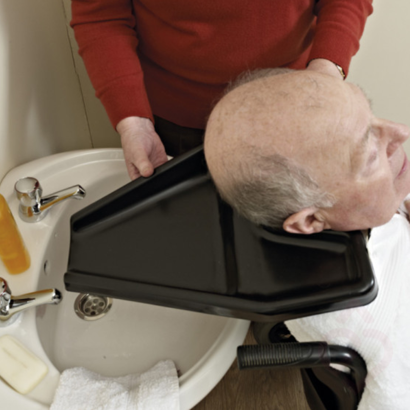 Pessoa com mobilidade reduzida a utilizar o lava-cabeças com auxílio de um cuidador, facilitando a lavagem do cabelo numa posição confortável sem necessidade de se inclinar sobre o lavatório.