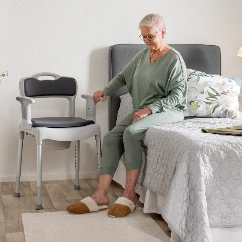 Cadeira sanitária e de duche regulável em altura, com braços de apoio, encosto e assento almofadado, colocada no quarto