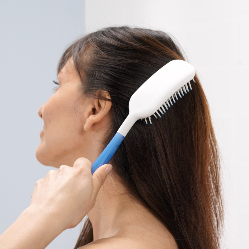 Mulher a utilizar a escova de cabelo de cabo longo, que permite alcançar o cabelo com maior facilidade e reduzir a necessidade de elevar o braço.