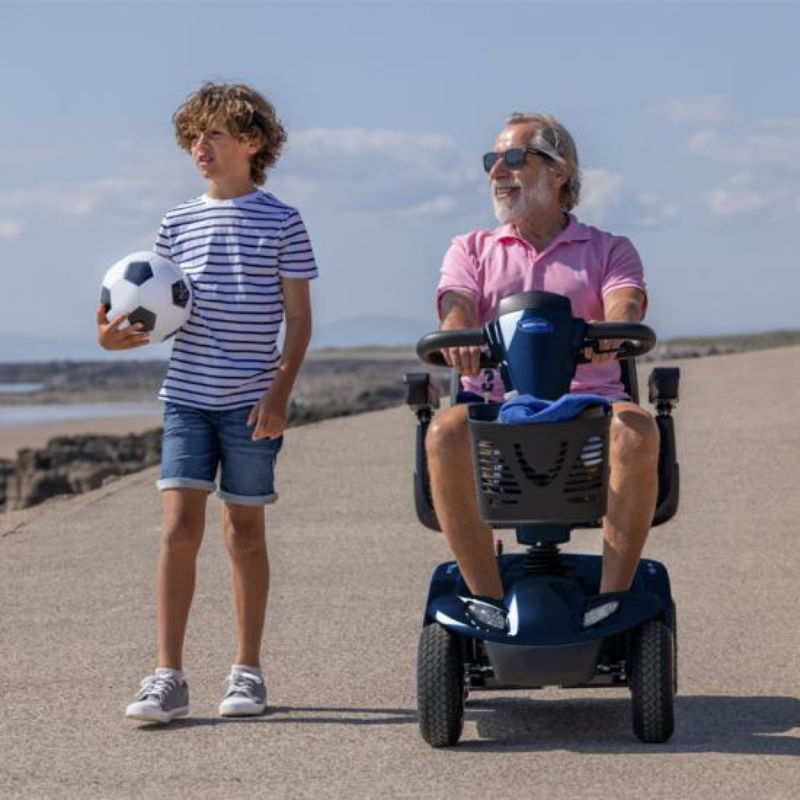 Homem a conduzir o scooter elétrico Invacare Leo, acompanhado por uma criança, num passeio junto à praia.